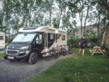An autumnal day where a motorhome parked in a wooded area, with three bikes resting against it