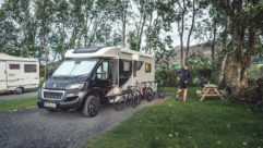 An autumnal day where a motorhome parked in a wooded area, with three bikes resting against it