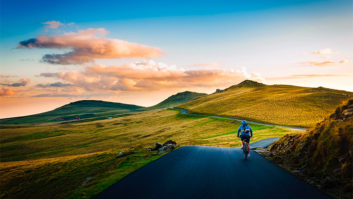 A person cycling along a path with green countryside all around and a sunset in the distance