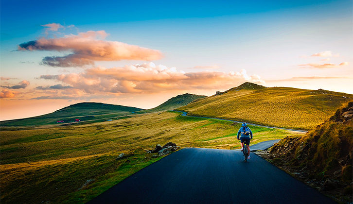 A person cycling along a path with green countryside all around and a sunset in the distance