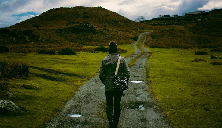A person walking along a wet gravel path with grass on either side and a hill in the distance on an overcast day