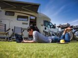 A woman laying on the grass on her laptop while her motorhome is in the background