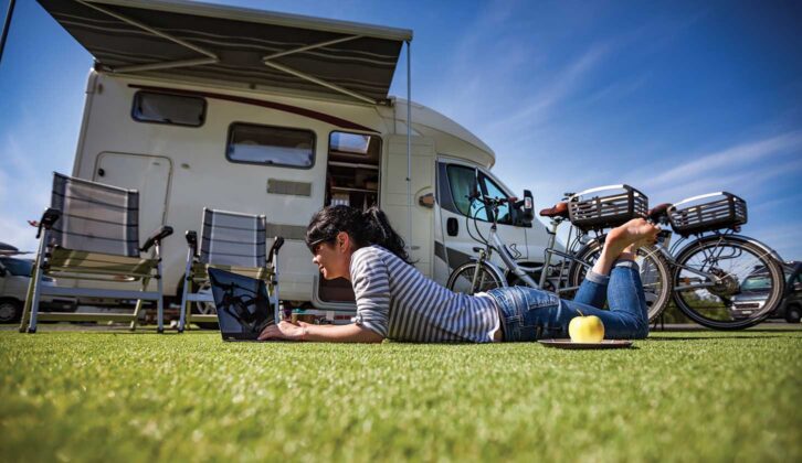 A woman laying on the grass on her laptop while her motorhome is in the background