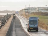 A campervan being driven down a road by the seafront
